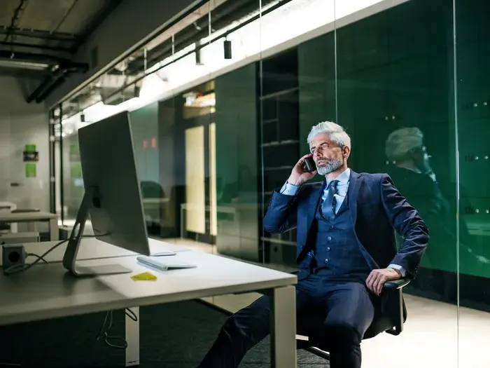 Mature businessman in a suit talks on the phone at a sleek desk in a glass walled office during late hours